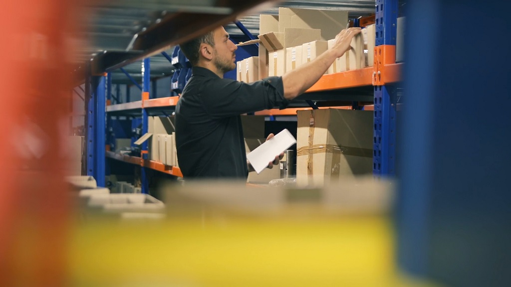 Warehouse employee organizing boxes on metal shelves while checking inventory with a tablet